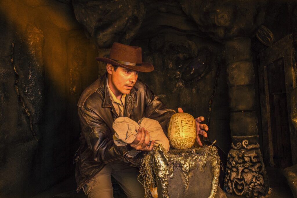 En la tenue luz de la cueva, un hombre con sombrero marrón y chaqueta de cuero busca un antiguo artefacto que recuerda a los tesoros de "Escape room Catacumbas en goldenpop". - ForTwo - Planes para parejas