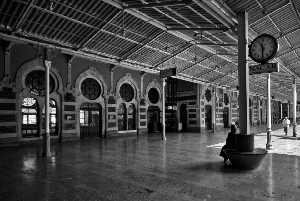 En una imagen en blanco y negro de una arquitectura ornamentada que evoca la grandeza de Zaragoza, una persona se sienta sola en una estación de tren vacía. Un gran reloj añade un toque de escapismo, similar a la atmósfera que se transmite en "Escapismo y Cluedo en Zaragoza". - ForTwo - Planes para parejas