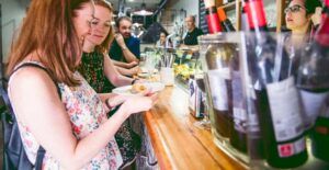 En el bullicioso Mercado de Mediodía y Tapas de Zaragoza, dos mujeres disfrutan de tapas en un animado bar, con copas de vino y botellas alineadas en la encimera, creando un ambiente enérgico perfecto para las delicias del mediodía. - ForTwo - Planes para parejas