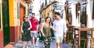 Cuatro amigos pasean y sonríen por una vibrante y estrecha calle de Zaragoza, donde las mesas rebosan de deliciosas tapas, capturando el espíritu animado del Mercado de Mediodía y Tapas en Zaragoza. - ForTwo - Planes para parejas