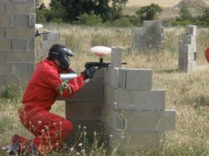 Vestido con un uniforme rojo, una persona se agacha detrás de un muro de hormigón, apuntando con una pistola de paintball mientras juega al Paintball en Murillo de Gállego (Zaragoza) en un campo al aire libre. - ForTwo - Planes para parejas