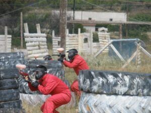 Dos individuos vestidos con monos rojos juegan al paintball, agazapados tras neumáticos y apuntando con sus marcadoras al aire libre en Paintball en Murillo de Gállego (Zaragoza). - ForTwo - Planes para parejas