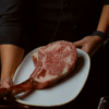 Una persona presenta un gran filete tomahawk marmolado en un plato blanco sobre un fondo oscuro, evocando la exquisita experiencia gastronómica en Parda BCN Una Cita con Encanto en Barcelona, donde cada plato se transforma en un momento inolvidable. - ForTwo - Planes para parejas