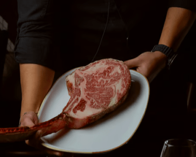 Una persona presenta un gran filete tomahawk marmolado en un plato blanco sobre un fondo oscuro, evocando la exquisita experiencia gastronómica en Parda BCN Una Cita con Encanto en Barcelona, donde cada plato se transforma en un momento inolvidable. - ForTwo - Planes para parejas