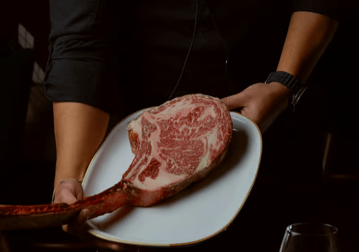 Una persona presenta un gran filete tomahawk marmolado en un plato blanco sobre un fondo oscuro, evocando la exquisita experiencia gastronómica en Parda BCN Una Cita con Encanto en Barcelona, donde cada plato se transforma en un momento inolvidable. - ForTwo - Planes para parejas