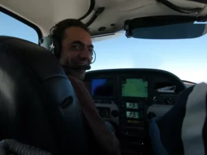 Persona sonriente en la cabina de una pequeña avioneta con auriculares, rodeada de instrumentos de navegación. A través de la ventana, los picos nevados sugieren el majestuoso Moncayo bajo un cielo despejado durante su paseo en avioneta de Zaragoza al Moncayo. - ForTwo - Planes para parejas