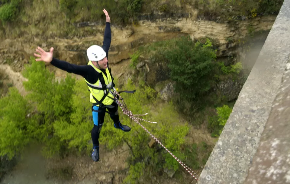 Un buscador de emociones se adentra en el Puenting en Murillo de Gállego, ataviado con casco y equipo de seguridad, y rodeado de un vibrante paisaje verde. - ForTwo - Planes para parejas