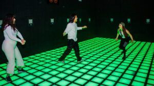 En una sala oscura, tres bailarines se mueven sincronizados sobre un suelo de rejilla verde brillante, electrificados por los ritmos de Pulse Up en Aventurico Barcelona, transformando el espacio con energía vibrante. - ForTwo - Planes para parejas