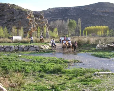 En la impresionante Sierra de Moncayo, jinetes a caballo y un perro recorren un arroyo poco profundo, enmarcado por acantilados rocosos y árboles: un día perfecto en la Ruta a Caballo por la Sierra de Moncayo. - ForTwo - Planes para parejas