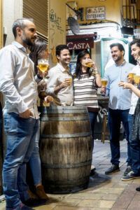 Un grupo de personas se reúne alrededor de una mesa de barril, con vasos de cerveza en mano, en una animada y decorada calle de Zaragoza. Es el escenario perfecto para su aventura del Tour de Tapas Nocturno en Zaragoza. - ForTwo - Planes para parejas