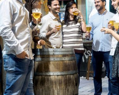 Un grupo de personas se reúne alrededor de una mesa de barril, con vasos de cerveza en mano, en una animada y decorada calle de Zaragoza. Es el escenario perfecto para su aventura del Tour de Tapas Nocturno en Zaragoza. - ForTwo - Planes para parejas