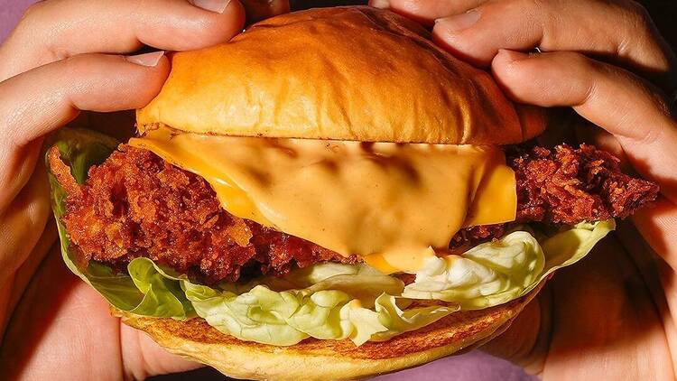Manos sosteniendo un delicioso sándwich de pollo con queso y lechuga en un pan, creando "Two Patties: El paraíso de las hamburguesas" para tu paladar. - ForTwo - Planes para parejas