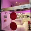 La entrada del restaurante cuenta con puertas de vidrio e iluminación rosa, con un atractivo cartel que dice "Two Patties: El paraíso de las hamburguesas" que conduce a un interior moderno perfecto para los amantes de las hamburguesas, completo con elegantes asientos de bar. - ForTwo - Planes para parejas