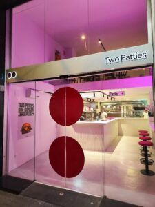 La entrada del restaurante cuenta con puertas de vidrio e iluminación rosa, con un atractivo cartel que dice "Two Patties: El paraíso de las hamburguesas" que conduce a un interior moderno perfecto para los amantes de las hamburguesas, completo con elegantes asientos de bar. - ForTwo - Planes para parejas