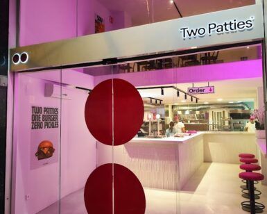 La entrada del restaurante cuenta con puertas de vidrio e iluminación rosa, con un atractivo cartel que dice "Two Patties: El paraíso de las hamburguesas" que conduce a un interior moderno perfecto para los amantes de las hamburguesas, completo con elegantes asientos de bar. - ForTwo - Planes para parejas