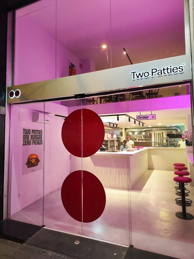La entrada del restaurante cuenta con puertas de vidrio e iluminación rosa, con un atractivo cartel que dice "Two Patties: El paraíso de las hamburguesas" que conduce a un interior moderno perfecto para los amantes de las hamburguesas, completo con elegantes asientos de bar. - ForTwo - Planes para parejas