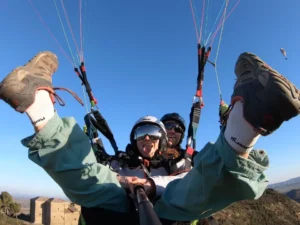Dos personas disfrutan de un vuelo en parapente en Lusía, sacando juguetonamente la lengua contra el cielo azul claro, con el impresionante paisaje extendiéndose debajo. - ForTwo - Planes para parejas