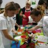 En Zaragoza, los participantes se ponen delantales y se reúnen alrededor de una mesa llena de utensilios de cocina, guiados por alguien con un delantal rojo durante el evento 👨‍🍳 Gymkana de MasterChef en Zaragoza: ¡Cocinad en pareja! 🍽️. - ForTwo - Planes para parejas