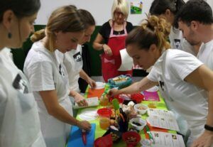 En Zaragoza, los participantes se ponen delantales y se reúnen alrededor de una mesa llena de utensilios de cocina, guiados por alguien con un delantal rojo durante el evento 👨‍🍳 Gymkana de MasterChef en Zaragoza: ¡Cocinad en pareja! 🍽️. - ForTwo - Planes para parejas