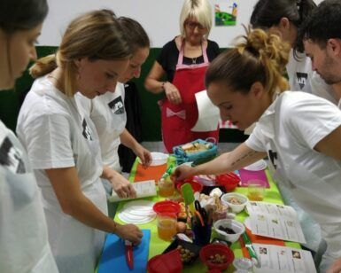 En Zaragoza, los participantes se ponen delantales y se reúnen alrededor de una mesa llena de utensilios de cocina, guiados por alguien con un delantal rojo durante el evento 👨‍🍳 Gymkana de MasterChef en Zaragoza: ¡Cocinad en pareja! 🍽️. - ForTwo - Planes para parejas