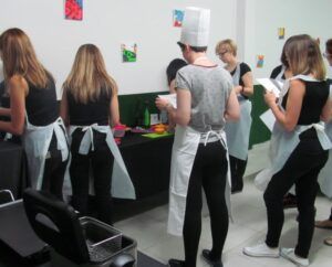En Zaragoza, la gente se pone delantales y gorros para la clase de cocina "👨‍🍳 Gymkana de MasterChef en Zaragoza: ¡Cocinad en pareja! 🍽️", reuniéndose alrededor de mesas llenas de ingredientes frescos. - ForTwo - Planes para parejas