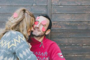Una pareja sonríe juguetonamente con pegatinas en forma de corazón en sus caras contra un fondo de madera, mostrando su conexión emocional. - ForTwo - Planes para parejas