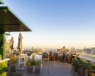 B-Heaven Rooftop Barcelona ofrece una experiencia elegante con sillas, mesas y grandes letras iluminadas que forman la palabra VIEW, todo ello perfectamente ambientado con un impresionante paisaje urbano al atardecer. - ForTwo - Planes para parejas