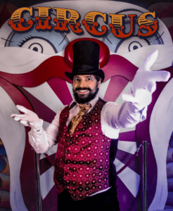 Un extraño artista de circo con chaleco y sombrero de copa se sitúa delante de un fondo con una cara de payaso con la palabra "CIRCUS" claramente encima, invitándote a vivir la experiencia Bizarre Escape Room Barcelona. - ForTwo - Planes para parejas