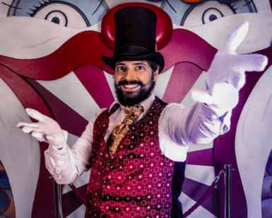 Un extraño artista de circo con chaleco y sombrero de copa se sitúa delante de un fondo con una cara de payaso con la palabra "CIRCUS" claramente encima, invitándote a vivir la experiencia Bizarre Escape Room Barcelona. - ForTwo - Planes para parejas