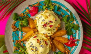 El "Brunch más original en Barcelona" está bellamente ambientado con un vibrante fondo rosa, presentando un llamativo plato de huevos Benedict adornado con flores comestibles y patatas. - ForTwo - Planes para parejas