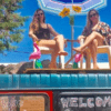 Dos adultos sentados en el techo de una furgoneta con sillas, sombrilla e hinchables, capturan a la perfección el ambiente despreocupado de un campamento para adultos en Ibiza. El cielo azul y los árboles realzan la tranquilidad del lugar. - ForTwo - Planes para parejas
