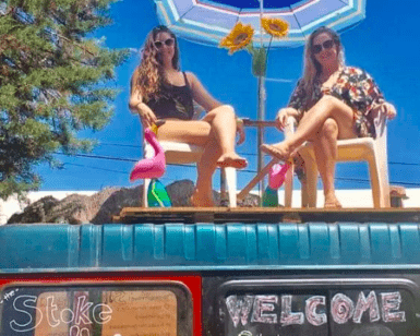Dos adultos sentados en el techo de una furgoneta con sillas, sombrilla e hinchables, capturan a la perfección el ambiente despreocupado de un campamento para adultos en Ibiza. El cielo azul y los árboles realzan la tranquilidad del lugar. - ForTwo - Planes para parejas