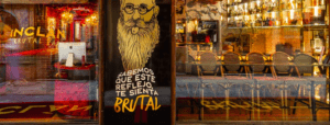 La vista de la calle de Inclán Brutal Bar muestra una ilustración de un hombre barbudo con texto en español en la fachada de vidrio, promoviendo la "Cena creativa con espectáculo en Inclán Brutal Bar" para una experiencia de sabores diversos y una cena creativa. - ForTwo - Planes para parejas