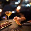 Una persona sostiene comida en una mesa poco iluminada con bebidas y aperitivos; las luces borrosas de la ciudad y las vistas panorámicas de Barcelona crean un telón de fondo de ensueño, ideal para una cena romántica con vistas panorámicas en Barcelona. - ForTwo - Planes para parejas