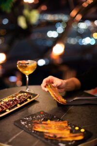 Una persona sostiene comida en una mesa poco iluminada con bebidas y aperitivos; las luces borrosas de la ciudad y las vistas panorámicas de Barcelona crean un telón de fondo de ensueño, ideal para una cena romántica con vistas panorámicas en Barcelona. - ForTwo - Planes para parejas