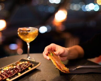 Una persona sostiene comida en una mesa poco iluminada con bebidas y aperitivos; las luces borrosas de la ciudad y las vistas panorámicas de Barcelona crean un telón de fondo de ensueño, ideal para una cena romántica con vistas panorámicas en Barcelona. - ForTwo - Planes para parejas
