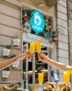 Dos parejas amantes de la comida brindan con mimosas afuera de una encantadora panadería de brunch con decoración floral, que refleja el ambiente vibrante de EatMyTrip Barcelona: Un lugar original para parejas amantes de la comida. - ForTwo - Planes para parejas