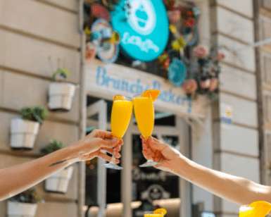 Dos parejas amantes de la comida brindan con mimosas afuera de una encantadora panadería de brunch con decoración floral, que refleja el ambiente vibrante de EatMyTrip Barcelona: Un lugar original para parejas amantes de la comida. - ForTwo - Planes para parejas
