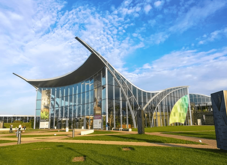 En Bucarest, un moderno edificio de cristal con techo curvo y pancartas se alza majestuoso bajo un cielo azul entre la exuberante vegetación. Esta joya arquitectónica le invita a vivir la experiencia de "Escapada a Bucarest y al spa más grande de Europa" con su elegante diseño. - ForTwo - Planes para parejas