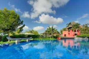 Relájese junto a la tranquila piscina de Escapada exótica para parejas en Alicante, donde una gran piscina azul refleja una vegetación vibrante y un llamativo edificio rojo bajo un cielo despejado, perfecto para parejas que buscan serenidad. - ForTwo - Planes para parejas
