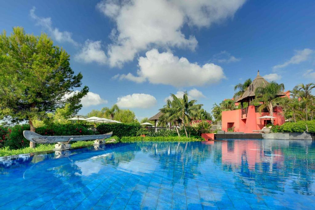 Relájese junto a la tranquila piscina de Escapada exótica para parejas en Alicante, donde una gran piscina azul refleja una vegetación vibrante y un llamativo edificio rojo bajo un cielo despejado, perfecto para parejas que buscan serenidad. - ForTwo - Planes para parejas
