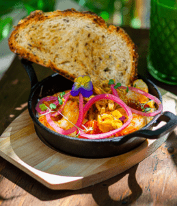 Experimenta el brunch artesanal en Lato Café Barcelona con camarones y vegetales en sartén, adornado con flores comestibles y servido sobre pan tostado sobre una tabla de madera rústica. - ForTwo - Planes para parejas