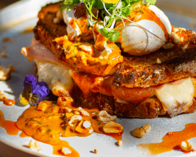 En Lato Café Barcelona, saborea un sándwich gourmet con huevos escalfados, verduras, salsa picante sobre pan crujiente, adornado con flores comestibles y nueces para una experiencia de brunch artesanal perfecta. - ForTwo - Planes para parejas
