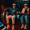 En el Museo del Videojuego de Madrid, dos personas se sumergen en un emocionante simulador de movimiento usando gafas de realidad virtual, con "EXPERIMENTA" mostrado debajo. - ForTwo - Planes para parejas