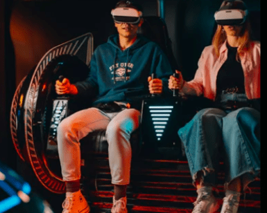 En el Museo del Videojuego de Madrid, dos personas se sumergen en un emocionante simulador de movimiento usando gafas de realidad virtual, con "EXPERIMENTA" mostrado debajo. - ForTwo - Planes para parejas