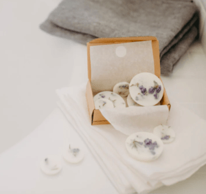 Tablillas de cera redondas con flores de color púrpura se muestran junto a una caja abierta, apoyada sobre telas blancas apiladas, que recuerdan a un taller de Barcelona conocido como Taller de velas de soja en Barcelona. - ForTwo - Planes para parejas