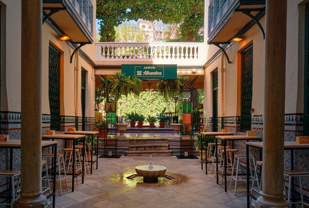 Esta cafetería con patio de Granada es un oasis con mesas y sillas, rodeado de exuberantes plantas y paredes de azulejos bajo el cartel que dice Jardín Alhambra. - ForTwo - Planes para parejas