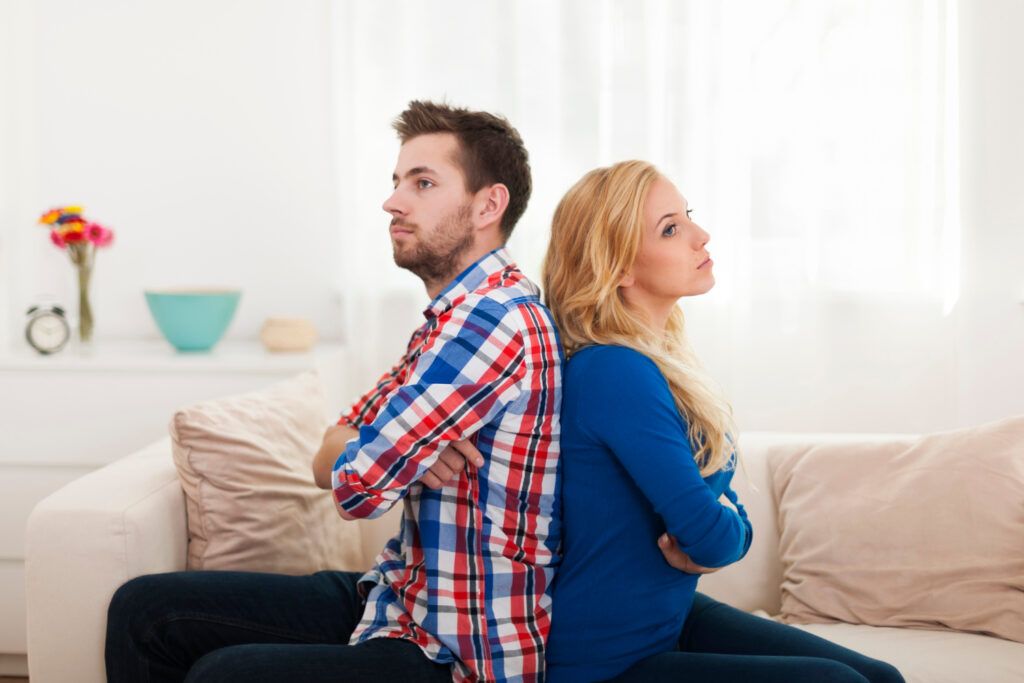 Hombre y mujer sentados espalda con espalda en un sofá, con los brazos cruzados, con aspecto frustrado después de otra instancia de discutir por tonterías. - ForTwo - Planes para parejas