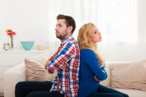 Hombre y mujer sentados espalda con espalda en un sofá, con los brazos cruzados, con aspecto frustrado después de otra instancia de discutir por tonterías. - ForTwo - Planes para parejas